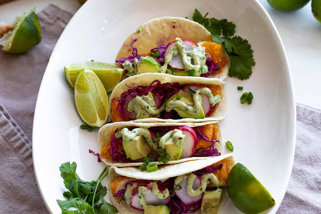 Plate of four vegan fish tacos topped with a creamy cilantro lime sauce made with dairy-free brie cheese.