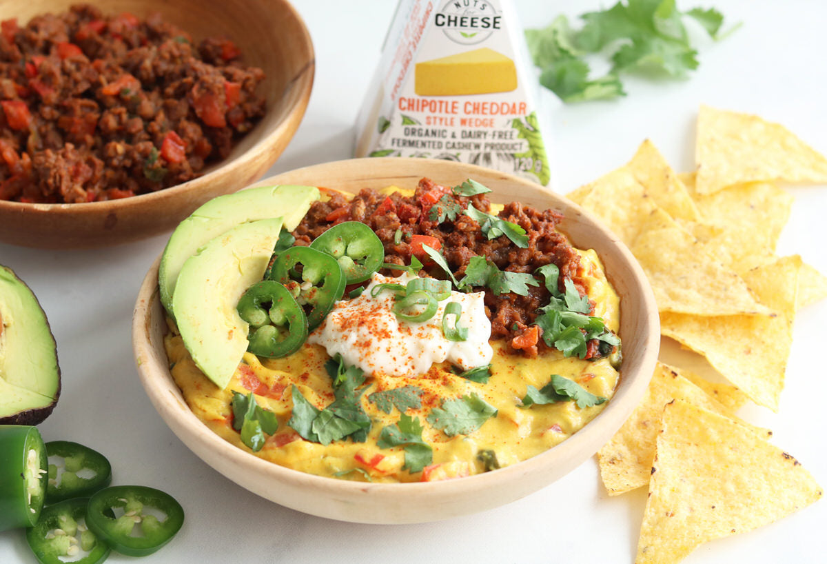 A bowl of vegan queso made from dairy-free chipotle cheddar cheese, topped with veggie ground, dairy-free sour cream, avocado, and jalapenos. Served with tortilla chips next to a box of dairy-free chipotle cheddar cheese.