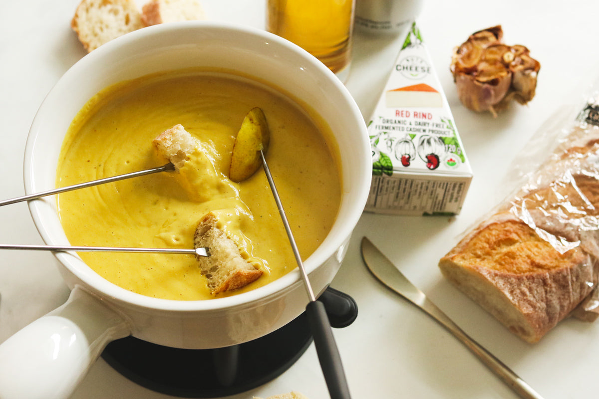 Fondue pot filled with melted dairy-free cheese. Three fondue forks with pieces of bread are seen dipping into the vegan cheese sauce. Next to the pot is a box of dairy-free smoky gouda cheese and a baguette.