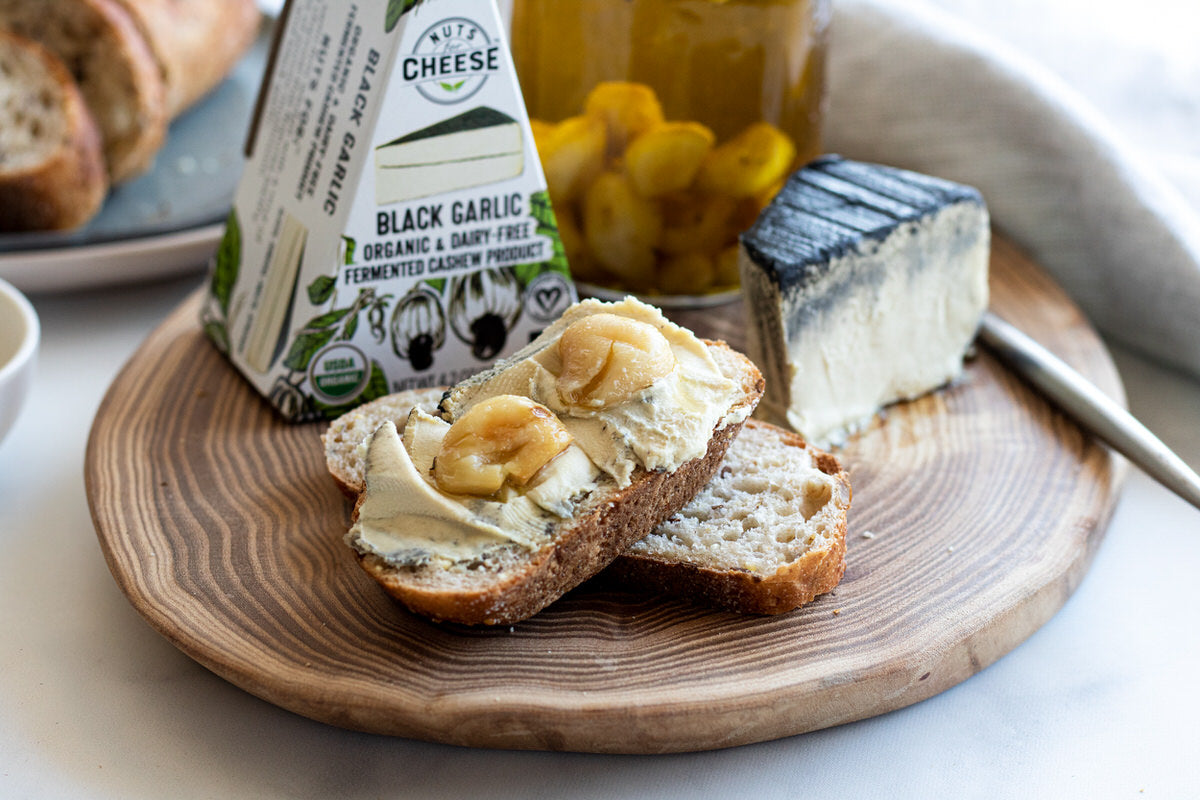 Two pieces of bread topped with dairy-free cheese and garlic confit sit on a wood plate. Beside them is a wedge of dairy-free black garlic cheese. 