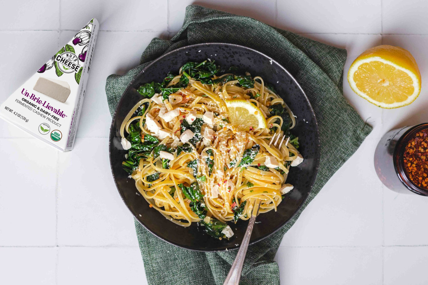 A pan full of prepared linguine with kale, set beside a box of dairy free brie cheese, a sliced lemon, and chili flakes.