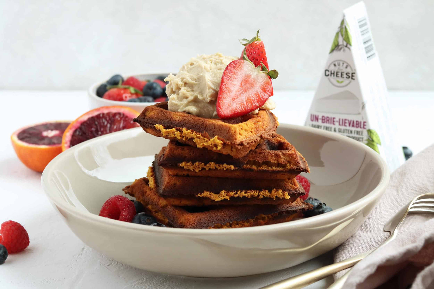 A stack of sweet potato cinnamon waffles in a bowl topped with dairy-free brie cheese and fresh strawberries. Served next to a box of dairy-free brie cheese.