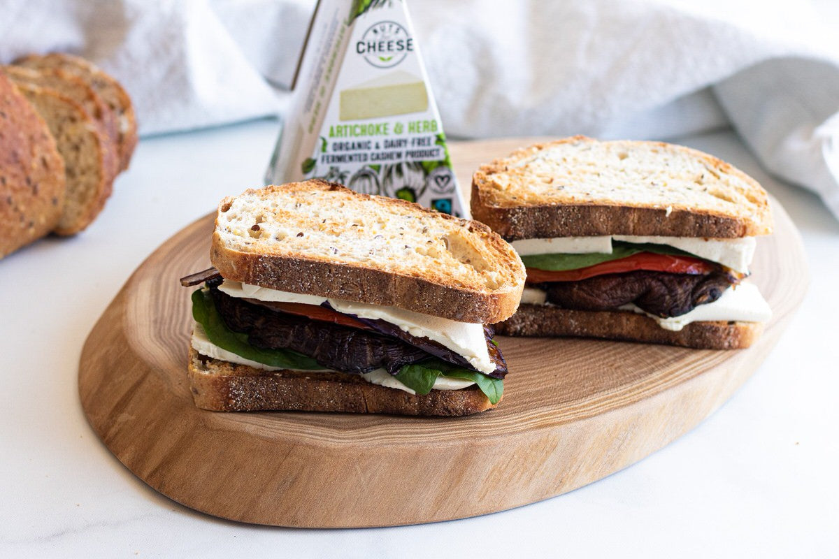 Two sandwiches made with grilled veggies and slices of dairy-free cheese sit on a wooden platter beside a box of dairy-free brie cheese.