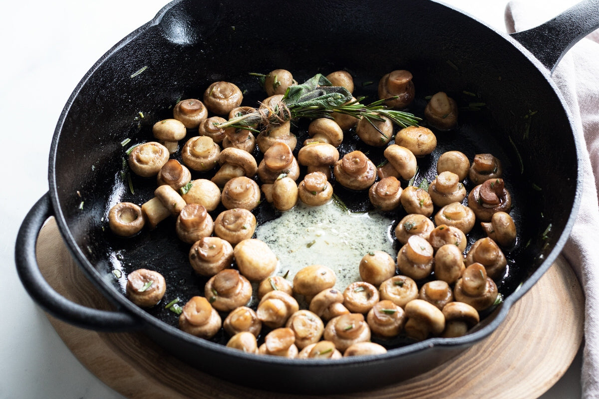 Frying pan filled with cooked mushrooms, melted dairy-free herb & garlic butter, and a bundle of fresh herbs.