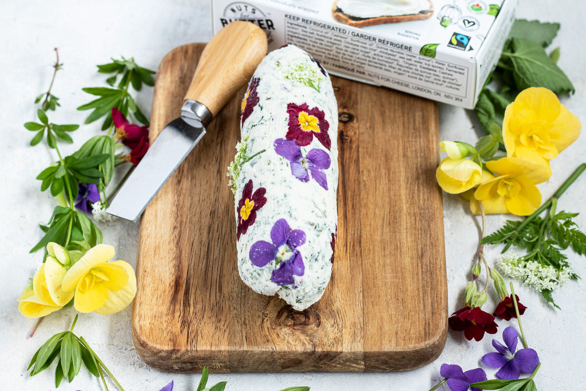 Log of dairy-free butter mixed with fresh herbs and flower petals, served on a wood cutting board with a butter knife. Surrounded by fresh flowers and a box of dairy-free butter.