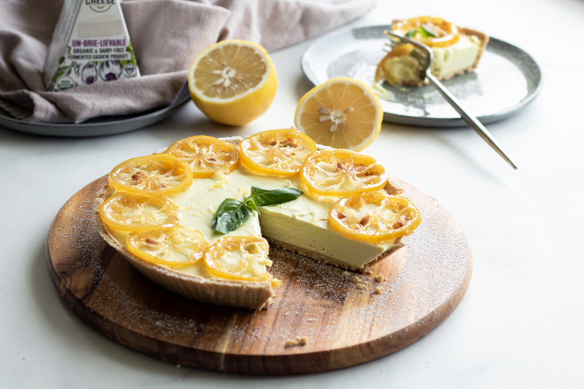 Prepared vegan lemon cheesecake topped with candied lemon slices, served on a wood platter. One slice has been cut from the cake and shown plated in the background with fresh lemons and a box of dairy-free brie cheese. 