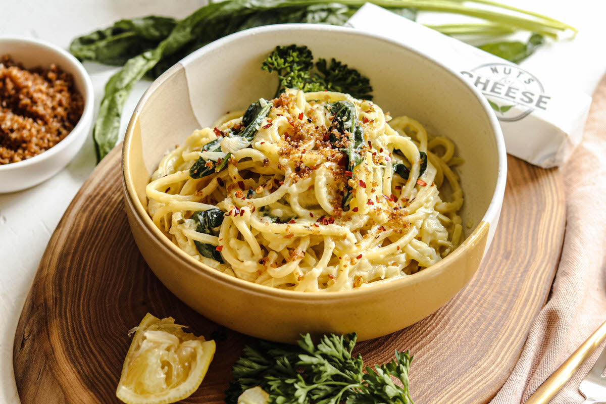 Bowl of cooked pasta with kale and a lemony sauce made with dairy-free artichoke & herb cheese. Topped with herbed breadcrumbs and served next to a stick of dairy-free butter. 