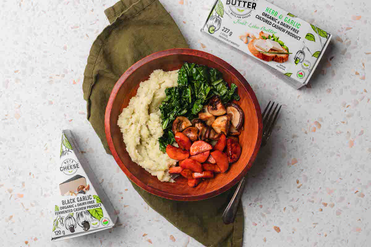 A bowl of dairy-free mashed potatoes topped with sauteed kale, mushrooms, and roasted carrots. Served next to boxes of dairy-free black garlic cheese and dairy-free butter.