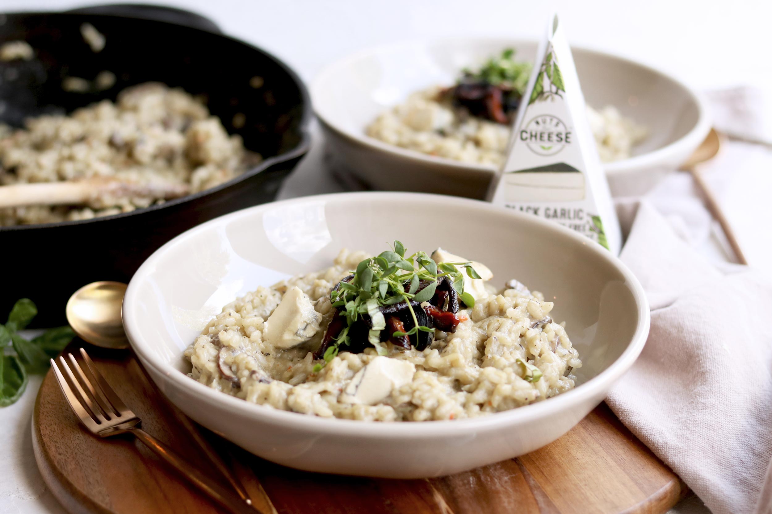 Two bowls of vegan mushroom risotto made with dairy-free black garlic cheese and topped with sauteed mushrooms. Served next to a pan with more risotto and a wedge of dairy-free black garlic cheese.