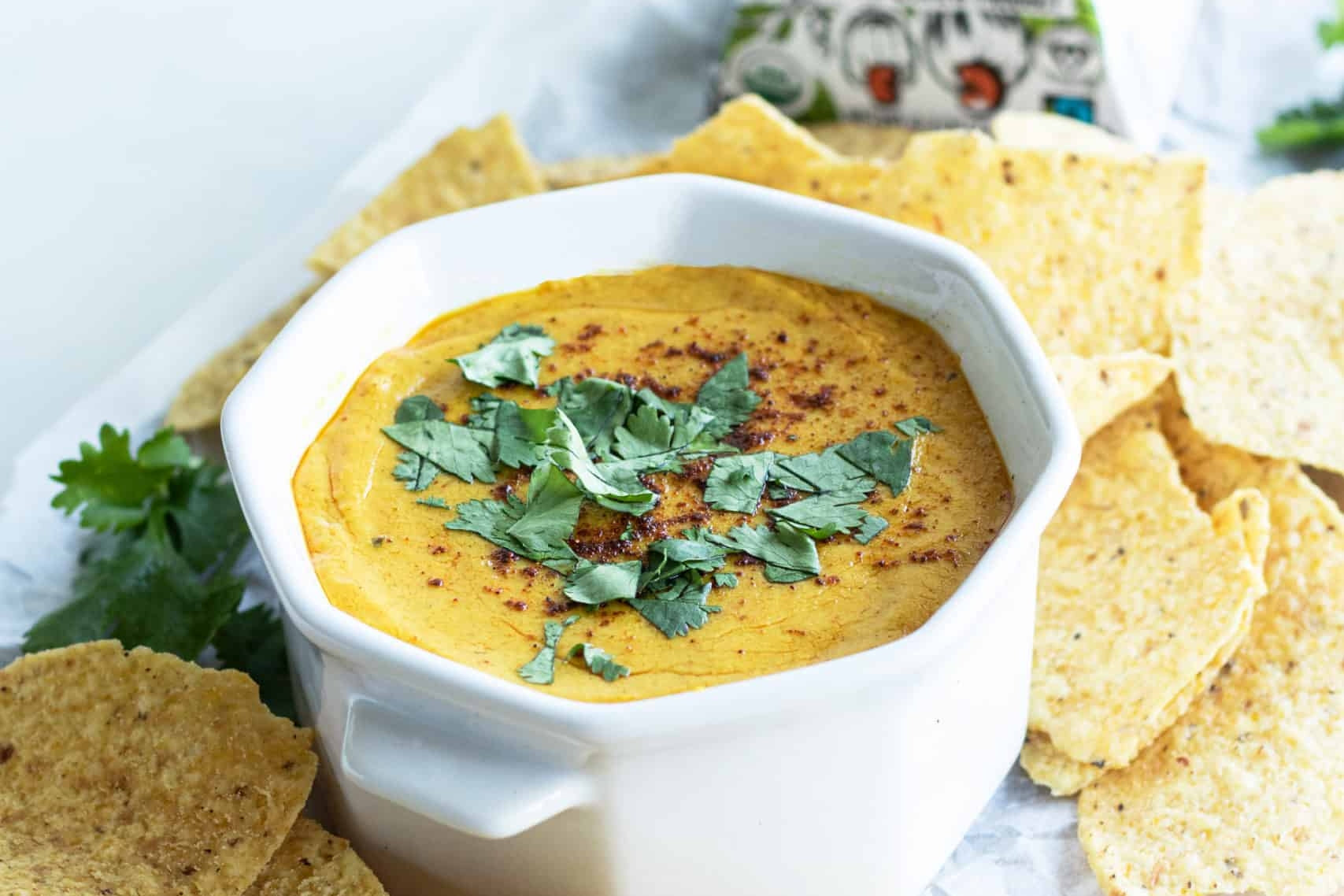 Bowl of vegan nacho cheese dip made with dairy-free chipotle cheddar cheese, topped with fresh cilantro and paprika. Served on a plate with tortilla chips and a box of dairy-free chipotle cheddar cheese.
