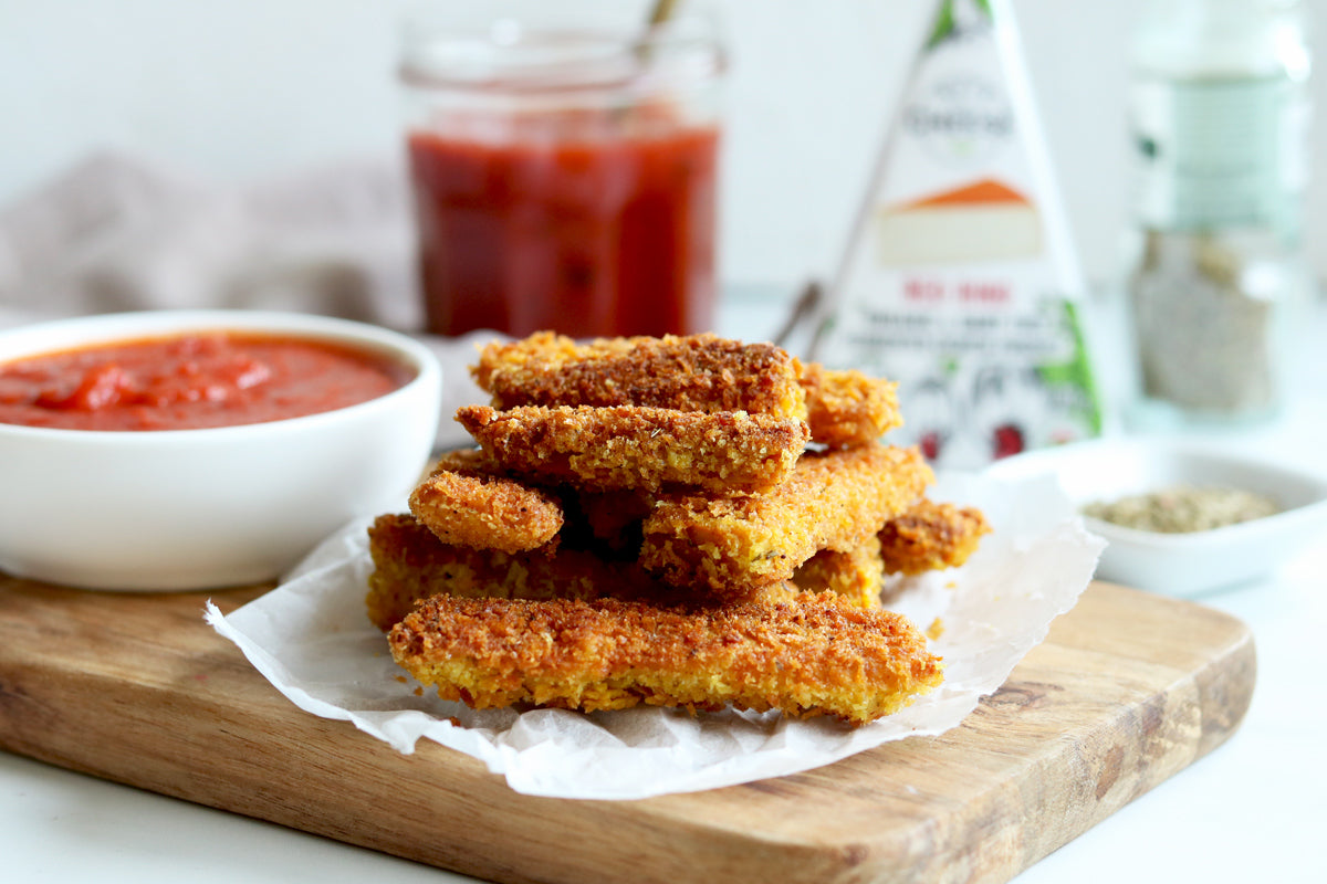 A stack of fried panko crusted vegan cheese sticks served with a bowl of marinara sauce beside a box of dairy-free smoky gouda cheese.