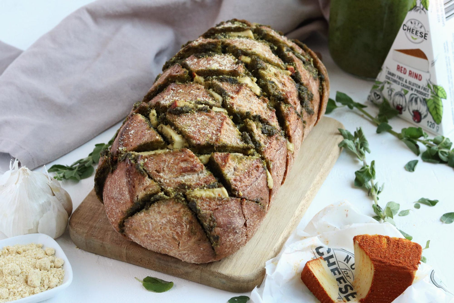 A loaf of bread stuffed with dairy-free gouda and pesto. Served on a cutting board next to a wedge of dairy-free smoky gouda cheese. 