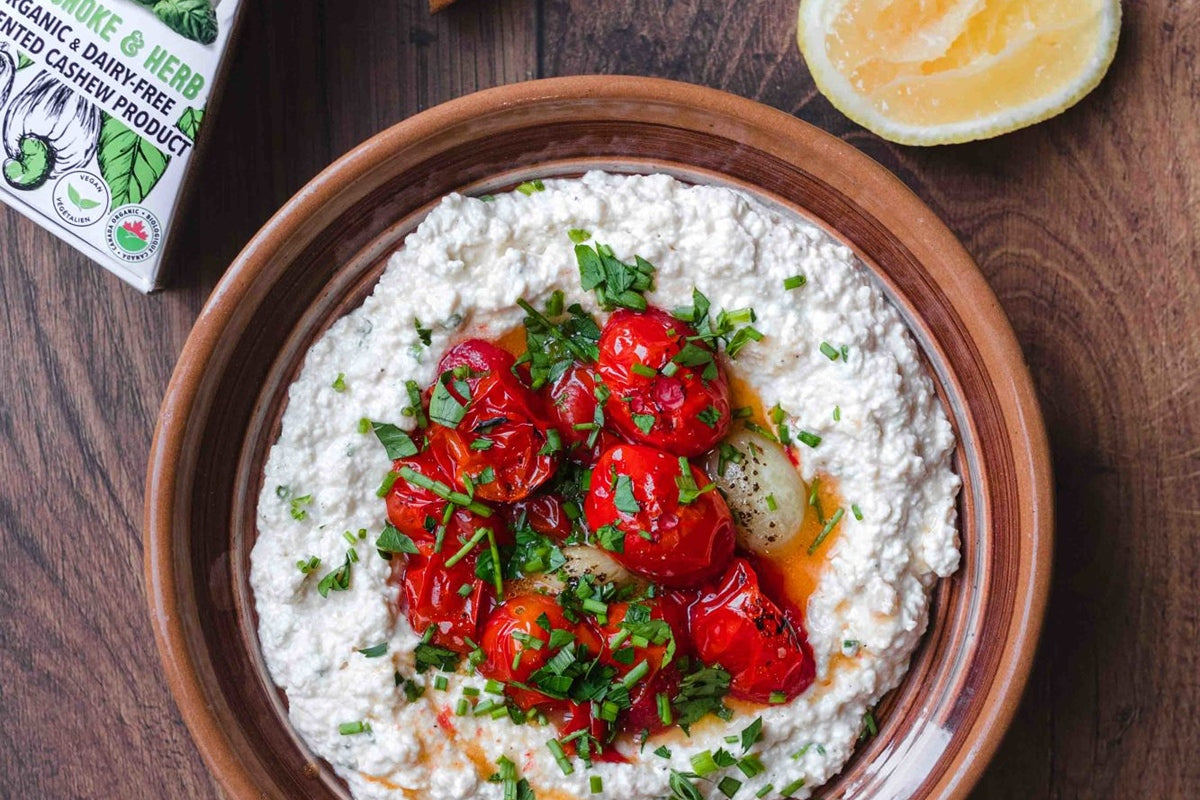Prepared bowl of dairy-free ricotta dip topped with roasted tomatoes and herbs. Set beside a slice of lemon and a box of dairy-free artichoke & herb cheese.
