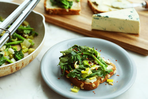 Slice of toast on a plate loaded with dairy-free blue cheese, rapini and leeks. Served next to a pan with more cooked rapini and leeks and a cutting board with a wedge of dairy free blue cheese.