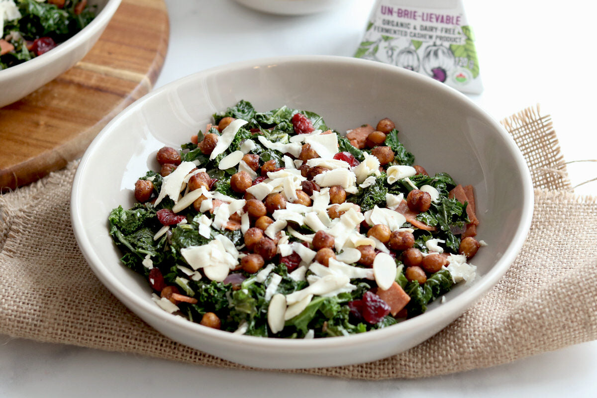 Large bowl of warm kale salad topped with roasted chickpeas, shredded dairy-free cheese, and drizzled with vegan bacon vinaigrette. Served next to a box of dairy-free brie cheese.