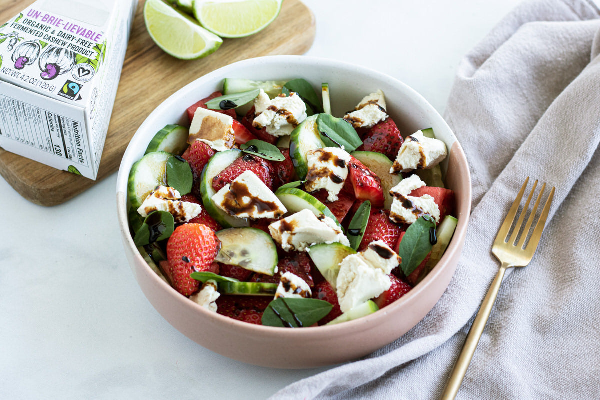 Bowl of prepared salad featuring cucumbers, strawberries, watermelon and chunks of dairy-free cheese drizzled with a balsamic dressing. Served next to a box of dairy-free brie and fresh lime wedges.