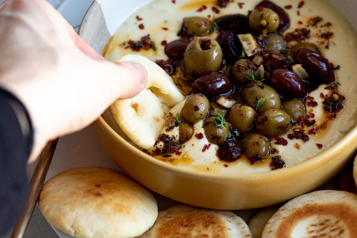 A hand is dipping bread into a bowl of dairy-free whipped brie dip which is topped with roasted olives. 
