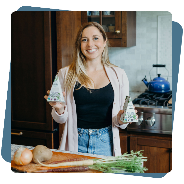 Margaret Coons, Nuts For Cheese™ Founder smiling while holding plant-based cheese products in the kitchen with vegetables on counter