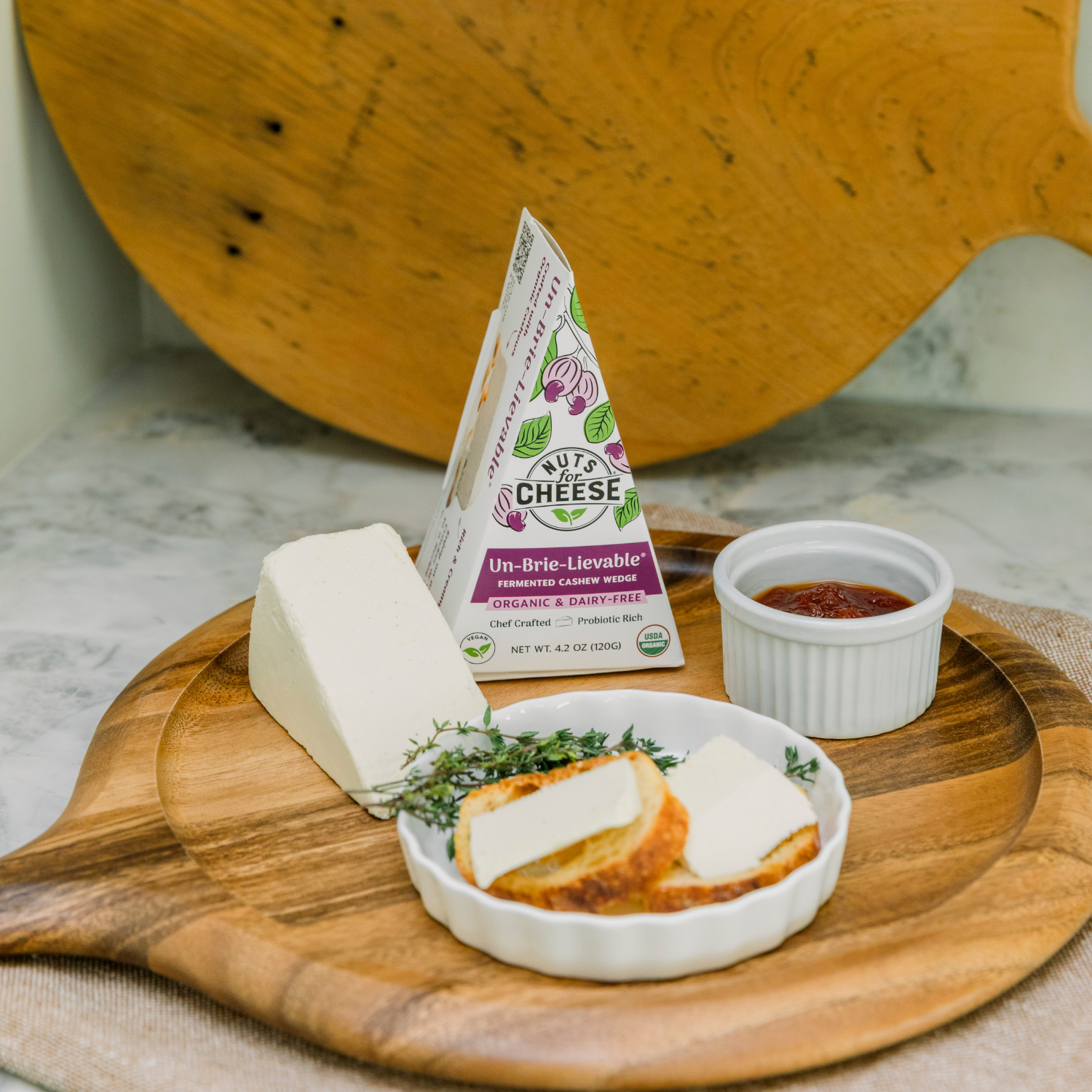 Planche à découper en bois avec un morceau de fromage, des tranches de pain et un petit récipient de sauce, avec un paquet de Nuts For Cheese en arrière-plan.