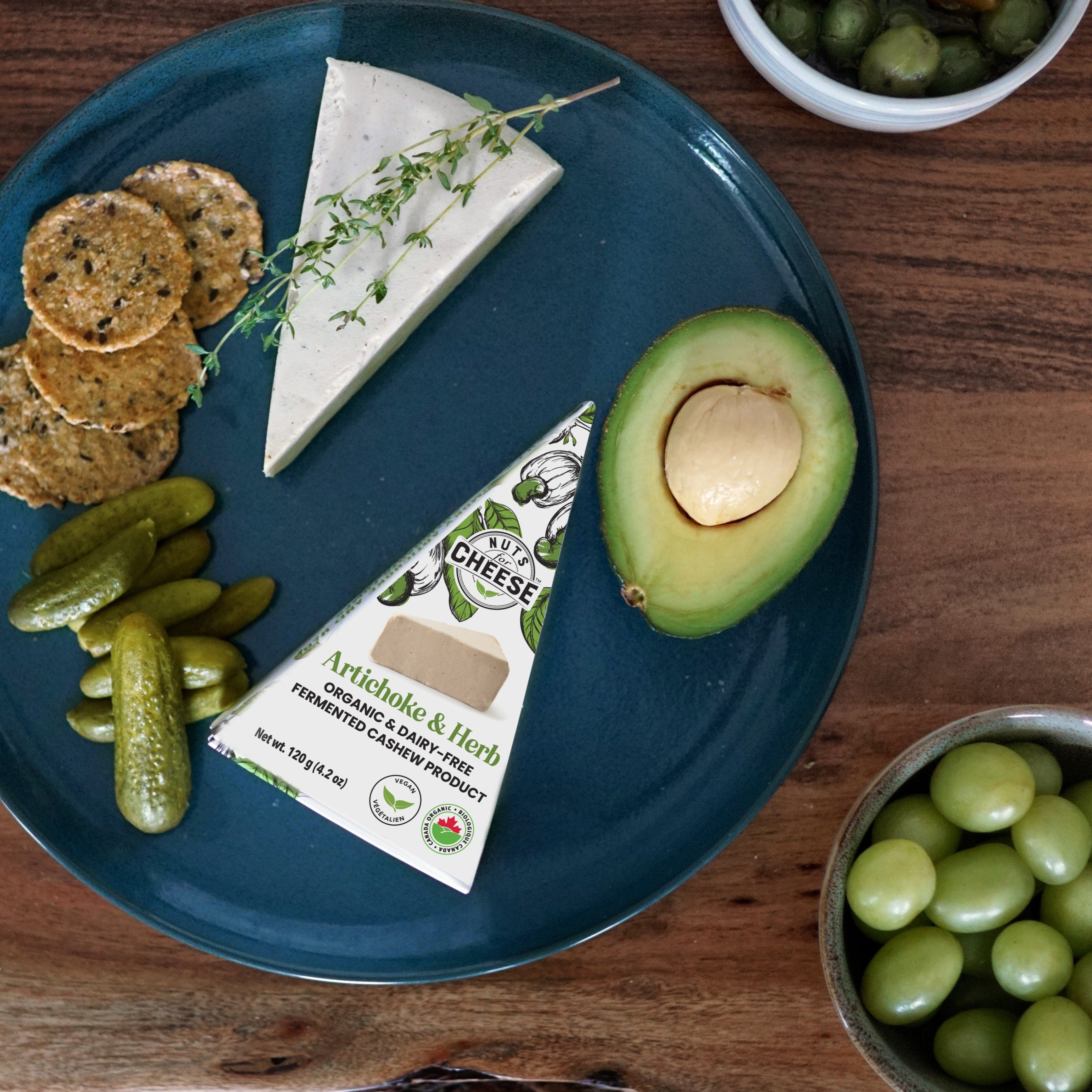Nuts For Cheese™ Organic & dairy-free fermented Artichoke & Herb cheese wedge plated on a wooden table with pickles, crackers, olives and avocado